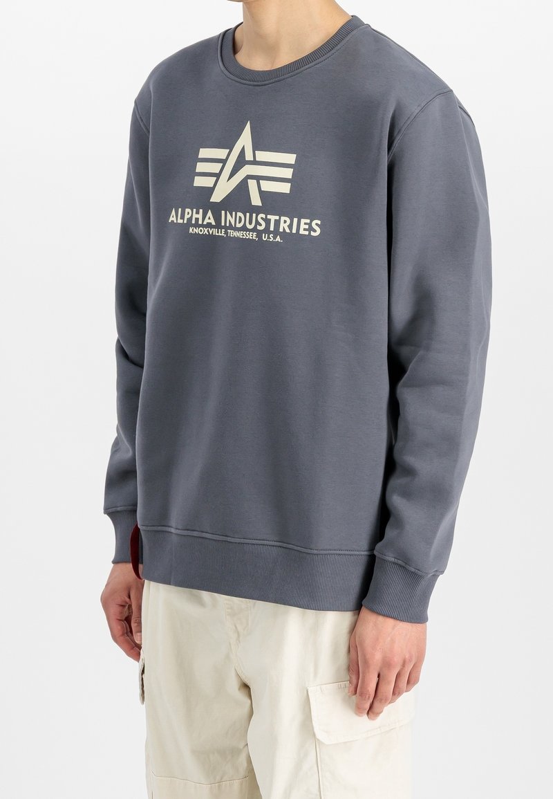 Sweat-shirt gris en tissu doux avec encolure ronde, poignets côtelés et un grand logo crème "ALPHA INDUSTRIES" sur le devant.
