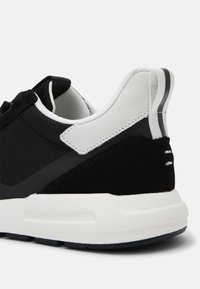 Chaussure de sport noire et blanche avec une tige en mesh, des accents en daim et une semelle en caoutchouc ; comporte un col rembourré et un détail rayé au talon.