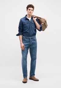 Tumši zils džinsu krekls ar pogām, kabatām, apvienots ar vidēji ziliem džinsiem. Brūni potītes zābaki un gaiši brūna soma uz pleca.