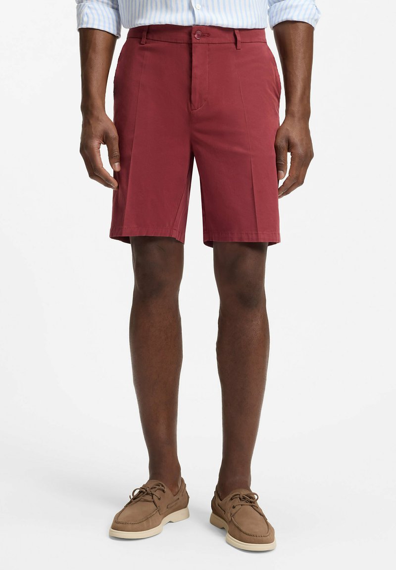 Homme portant un short bordeaux arrivant aux genoux, une chemise rayée bleu clair rentrée dans le short, et des mocassins beige, debout devant un fond blanc.
