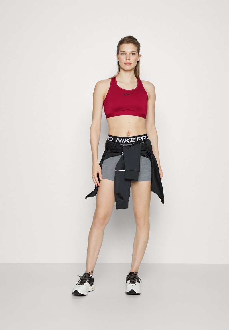 Reggiseno sportivo rosso con logo Nike nero, pantaloni corti grigi e una giacca nera legata in vita. Scarpe da ginnastica bianche con accenti scuri.