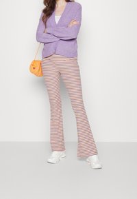 Lila stickad kofta över en vit topp, kombinerad med randiga utsvängda byxor med orange, blå och gula linjer; vita sneakers; orange väska.