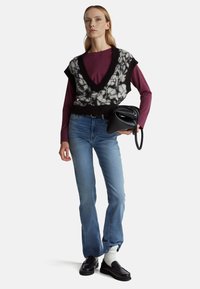 Pletená vesta v černé a bílé barvě přes vínovou košili s dlouhým rukávem, doplněná modrými džíny a černými mokasíny. Drží černou kabelku.