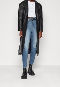 Černý kožený dlouhý kabát přes bílý strukturovaný crop top, spárovaný se světle modrými džíny a černými masivními kotníkovými botami; ladí s černou kabelkou.