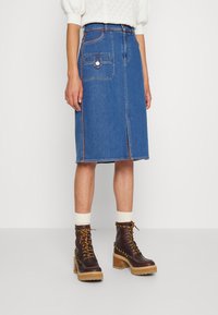 Mujer vistiendo una falda vaquera azul hasta la rodilla con abertura frontal, un top de punto color crema, calcetines blancos y botas de tacón grueso marrones con cordones.