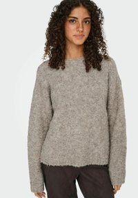 Jeune femme aux cheveux bouclés portant un pull en laine grise et un pantalon sombre, se tenant devant un fond clair et uni.