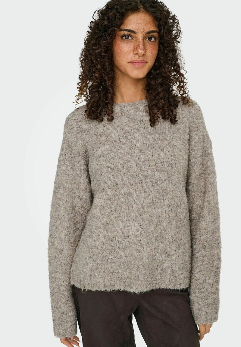 Jeune femme aux cheveux bouclés portant un pull en laine grise et un pantalon sombre, se tenant devant un fond clair et uni.