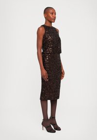 Abito con paillettes di un marrone scuro, design senza maniche, corpino aderente, gonna lunga fino al ginocchio. Abbinato a décolleté nere con cinturini e collant velati.