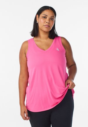 Frau trägt ein leuchtend rosa ärmelloses V-Ausschnitt-Oberteil und schwarze Hose, steht vor weißem Hintergrund und hält den Saum des Oberteils fest.