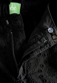 Pantalon noir avec des motifs floraux brodés, doté d'une fermeture éclair et d'un bouton, avec une étiquette verte portant la mention "XX". Tissu au toucher doux.