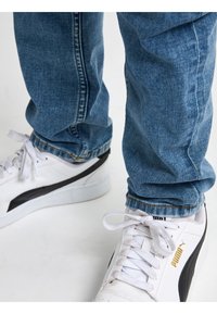 Baskets blanches Puma avec bande noire portées avec un jean bleu sur fond blanc.
