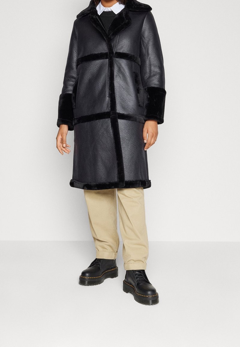 Abrigo de cuero negro con acentos de piel sintética, que presenta un cuello con solapa y bolsillos laterales. Se combina con pantalones beige y botas negras con cordones.