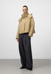 Beżowa kurtka cropped z wysokim kołnierzem, zapięciem na zatrzaski i dwiema przednimi kieszeniami, połączona z luźnymi, ciemnoszarymi spodniami i czarnymi butami.