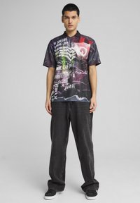 Polo de manga corta, negro y multicolor con gráficos que presentan varios textos y patrones. Combinado con jeans oscuros de corte holgado y zapatillas.