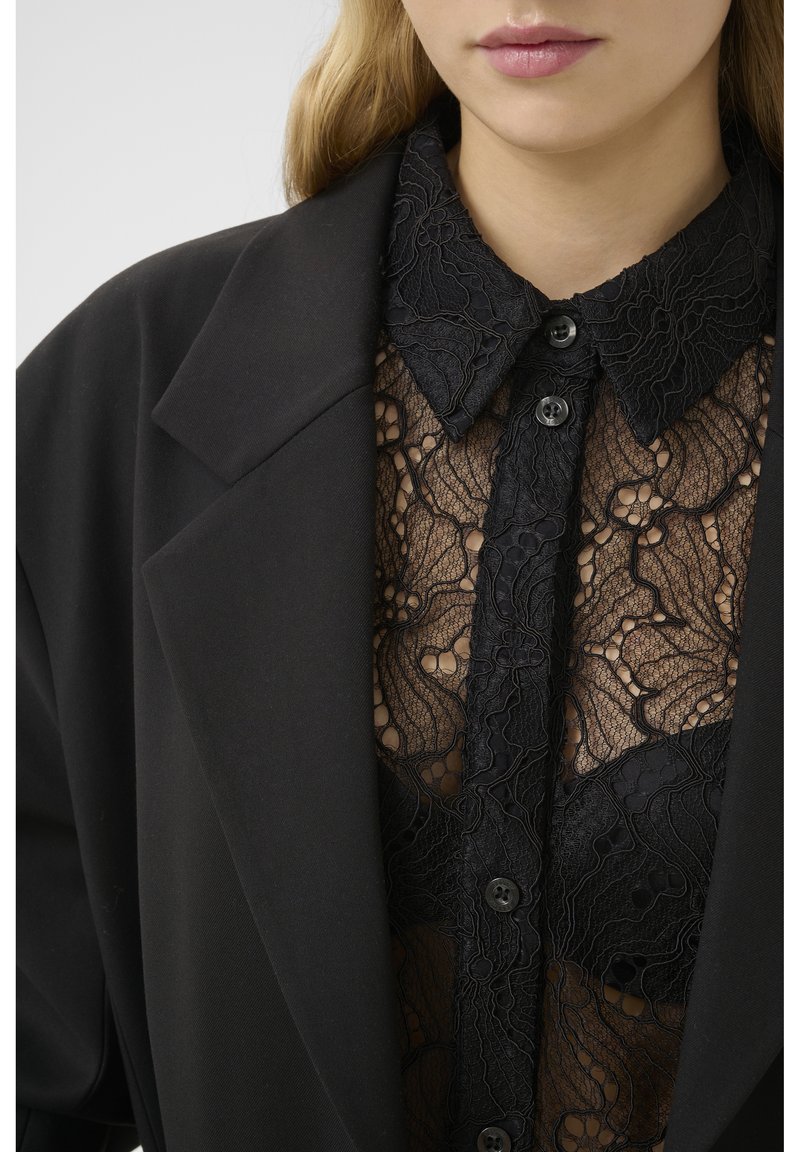 Schwarzer Blazer mit großen Revers, über einem durchsichtigen schwarzen Spitzenoberteil mit floralen Mustern und Knopfverschluss getragen. Schlanker Schnitt und passgenaues Design.