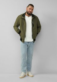 Olijf groene bomberjack met een rits aan de voorkant, zwarte kraag en manchetten, gedragen over een wit shirt, gecombineerd met lichtblauwe jeans en witte sneakers.