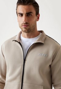Giacca beige con zip realizzata in morbido tessuto, dotata di un alto colletto. Presenta un logo grigio "GUESS" sul davanti e inserti neri sui lati.