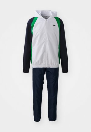 Giacca con zip, corpo bianco e maniche verde e blu navy; pantaloni blu navy abbinati. Tessuto leggero e liscio, design con cappuccio, polsini elasticizzati.