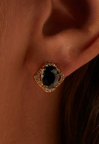 Goudkleurige oorbellen met een grote, facettaire zwarte steen omringd door kleine heldere stenen, gezet in een decoratief, geometrisch ontwerp.