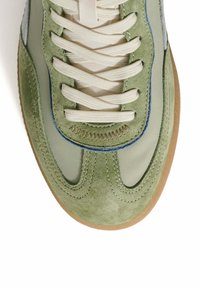 Zapatilla de ante verde con parte superior de cuero suave, cordones blancos y suela de goma marrón. Presenta acentos bordados y detalles azules en las costuras.
