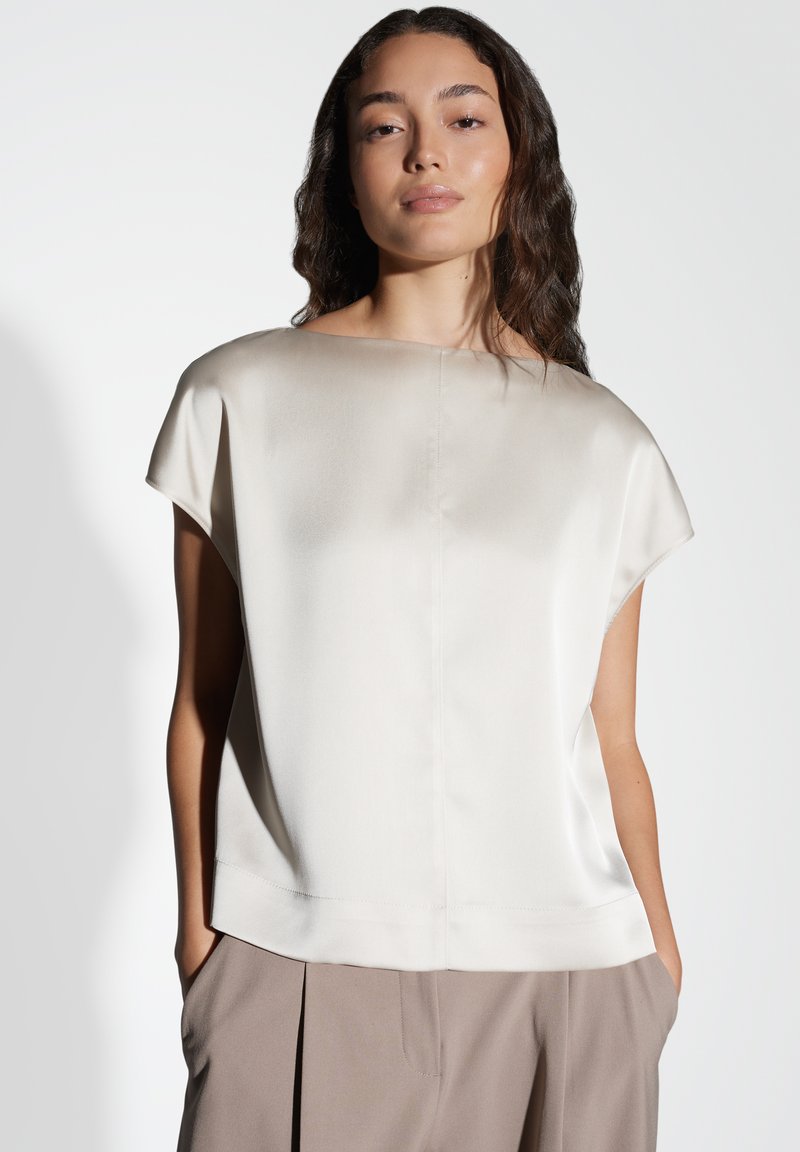 Top in seta crema a maniche corte con scollo a barca e morbido drappeggio. Abbinato a pantaloni color taupe, con una texture liscia e vestibilità rilassata.
