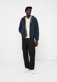 Navy hooded jacket with front zipper, paired with a light yellow polo and black cargo pants. Model wears a black cap and white sneakers.