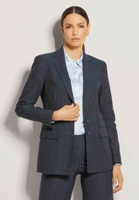 Marineblauer Nadelstreifenblazer mit zwei Knöpfen, Reverskragen und zwei Vorderaschen, getragen über einem hellblauen gemusterten Hemd.