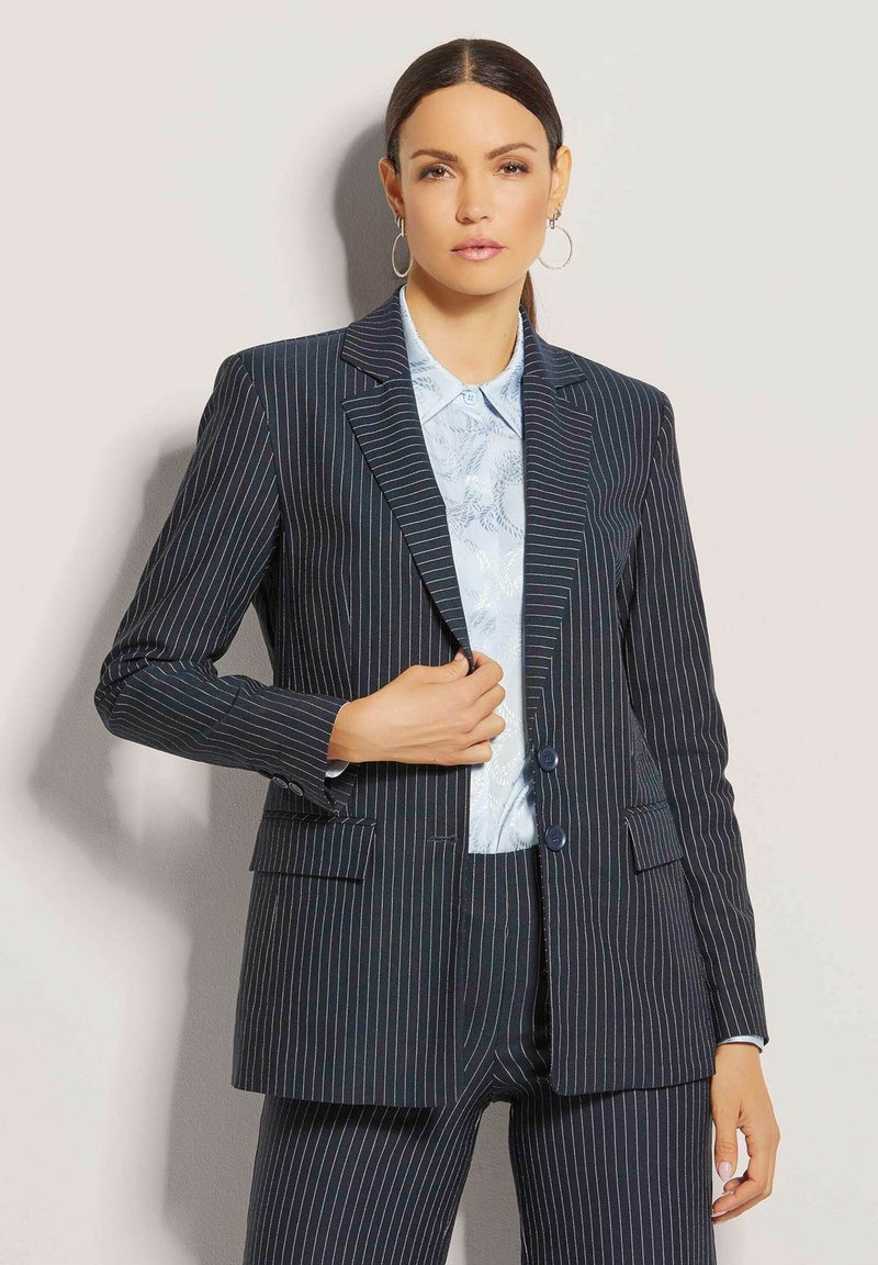Marineblauer Nadelstreifenblazer mit zwei Knöpfen, Reverskragen und zwei Vorderaschen, getragen über einem hellblauen gemusterten Hemd.