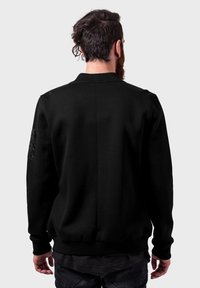 Svart genser med ribbestrikket krage, mansjetter og kant. Har en teksturert lapp på venstre erme. Minimalistisk design, behagelig passform.