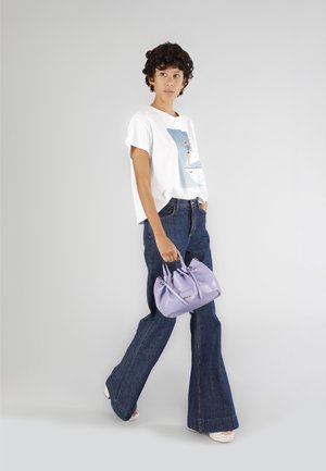 Witte grafische t-shirt, blauwe denim flared broek en een kleine lavendelkleurige handtas met dubbele handvatten. Model staat voor een neutrale achtergrond.