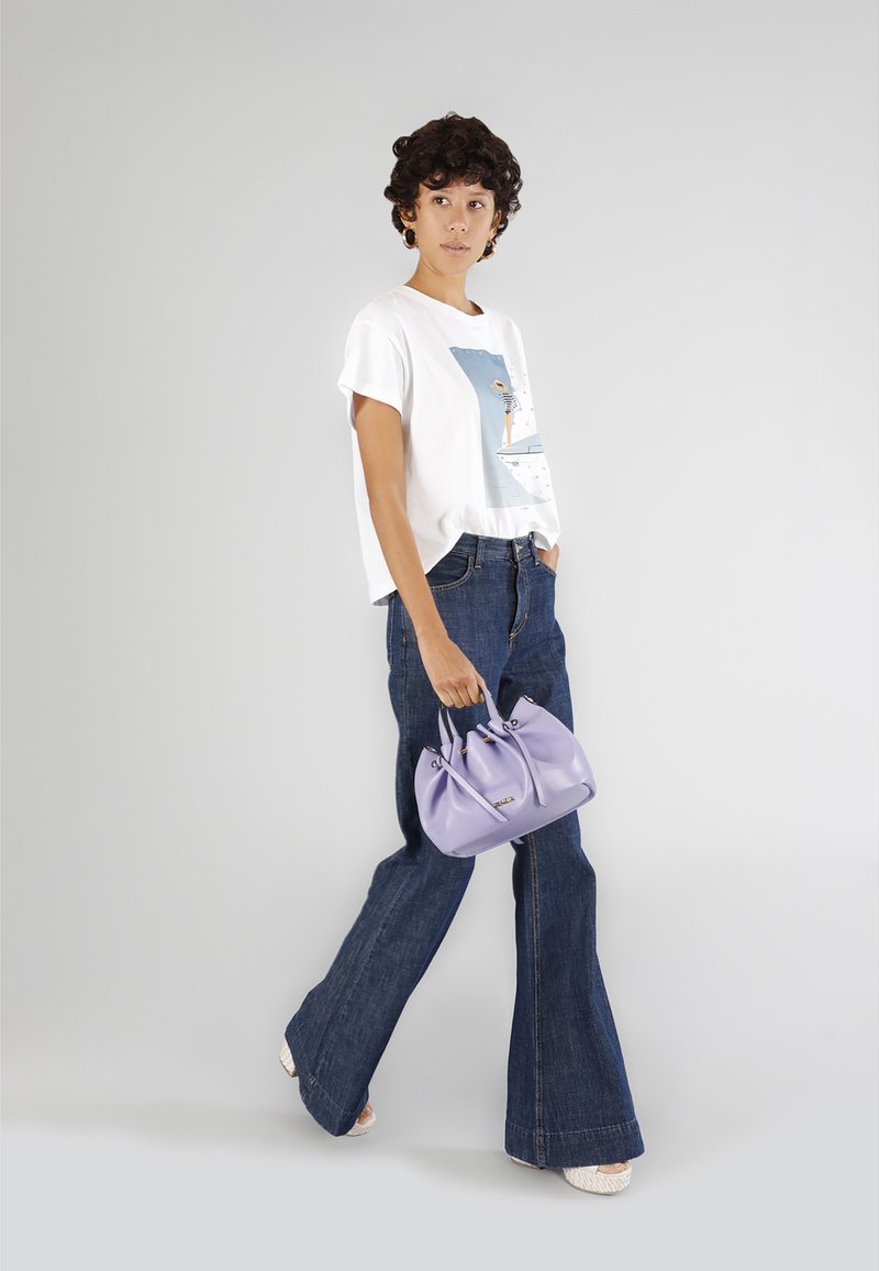 T-shirt grafica bianca, pantaloni in denim blu a campana e una piccola borsa lilla con manici doppi. La modella si trova su uno sfondo neutro.