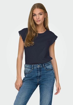 Joven mujer con cabello largo y rubio, vistiendo una blusa sin mangas de encaje en color navy y jeans de cintura alta azul, posando contra un fondo liso.