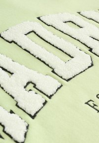 Tessuto verde chiaro con lettering bianco soffice contornato di nero. Il dettaglio della superficie testurizzata aggiunge profondità, con un design pulito e moderno.