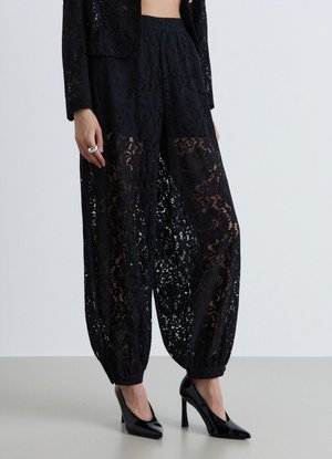 Donna che indossa pantaloni in pizzo nero trasparente con polsini elastici abbinati a scarpe nere con tacco alto e una giacca in pizzo coordinata.