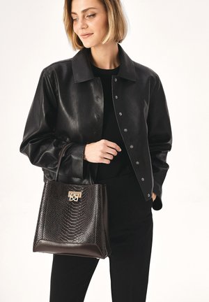 Veste en cuir noir avec boutons-pression portée sur un top noir, tenant un sac texturé marron foncé avec un logo doré.