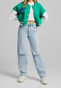 Giacca varsity verde e bianca con un design cropped, caratterizzata da ricami blu. Abbinata con jeans chiari strappati e sneaker bianche.