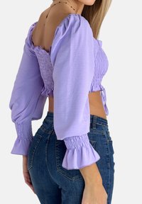 Lila gerafftes kurz geschnittenes Oberteil mit Puffärmeln, gerafften Details und Bindeband in der Taille, kombiniert mit hoch taillierten blauen Jeans.