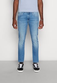 Man med ljusblå slim-fit jeans och svarta och vita sneakers, stående mot en enfärgad vit bakgrund.