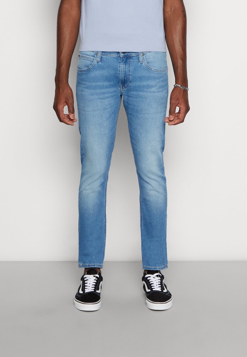 Man med ljusblå slim-fit jeans och svarta och vita sneakers, stående mot en enfärgad vit bakgrund.