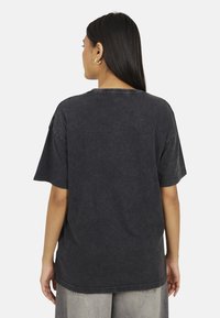 T-shirt nera oversize con maniche corte, scollo rotondo, texture sfumata e orlo grezzo. Indossato con pantaloni grigi a gamba larga.