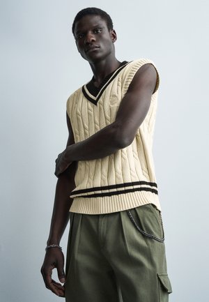 Młody mężczyzna ubrany w kremową dzianinową kamizelkę bez rękawów z czarnymi paskami oraz oliwkowe spodnie typu cargo z łańcuszkową ozdobą.