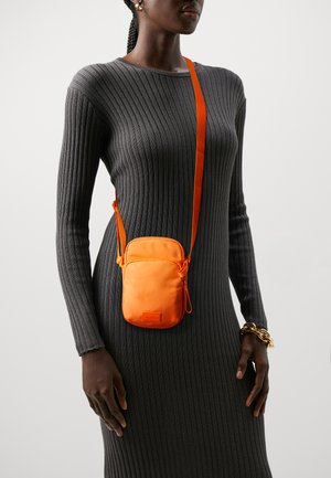 Sac banane en nylon orange avec un cordon de serrage, suspendu à une longue sangle. La personne qui le porte est dans une robe grise à côtes avec des manches longues.