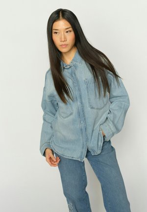 Kobieta o długich, prostych, czarnych włosach, ubrana w jasnoniebieską kurtkę dżinsową i niebieskie dżinsy, stojąca z jedną ręką w kieszeni.