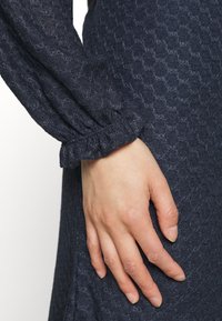 Vestido azul-marinho texturizado com padrão de colmeia; apresenta um punho franzido na manga, destacando um tecido suave e uma silhueta ajustada.