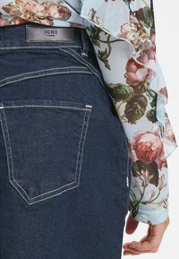 Mörkblå denimjeans har en bakficka med vit sömnad. En blommönstrad, genomskinlig blus är delvis synlig, med volangärmar.