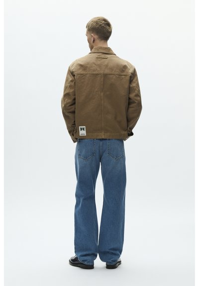 Ruskea canvas-takki, jossa on kaulus, takakappaleen yläosa ja logoplässi, yhdistetty vaaleansinisiin farkkuihin, joissa on klassinen viiden taskun malli.