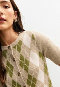 Donna che indossa un maglione a rombi beige e verde con bottoni, orecchini a cerchio dorati, su sfondo bianco semplice.