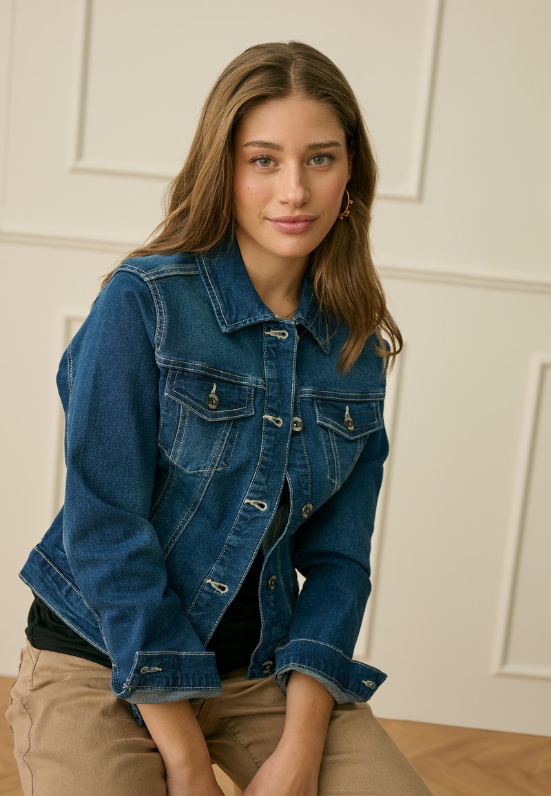 Joven con cabello largo, vestida con una chaqueta de mezclilla azul, pantalones color caqui y pendientes de aro dorados, sentada en el interior frente a una pared con paneles crema.