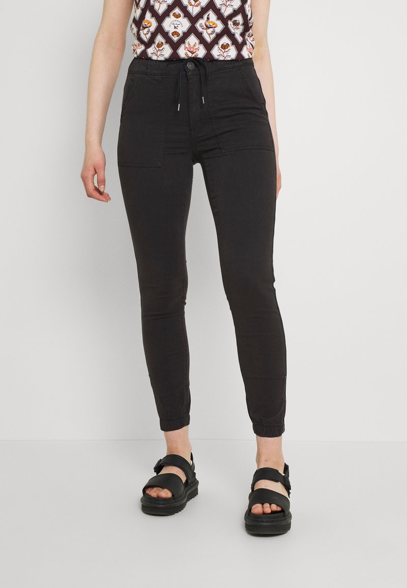 Femme portant un pantalon jogger noir avec des poches avant et des sandales plateformes noires, debout sur un fond blanc uni.