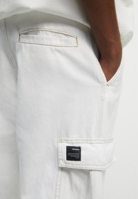 Pantalon en denim blanc avec poches cargo latérales, coutures contrastées et une étiquette noire de marque sur la poche. Texture lisse, coupe ajustée.